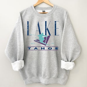 May include: A gray sweatshirt with the words "LAKE TAHOE" in a distressed navy blue font. A teal skier graphic is centered above the word "TAHOE". The sweatshirt is hanging on a wooden hanger.