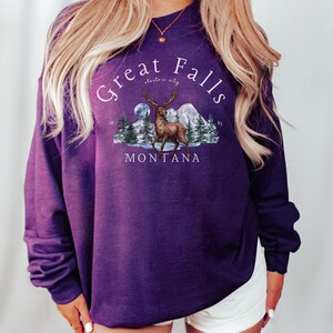 May include: Purple sweatshirt with a graphic print of a deer in a snowy mountain scene. The text "Great Falls electric city Montana" is printed on the sweatshirt.