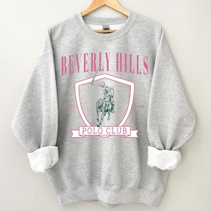 May include: Gray sweatshirt with a pink "Beverly Hills Polo Club" graphic featuring a polo player on a horse. The graphic is inside a shield with the years "Est'd 1990" and "1990" on either side.