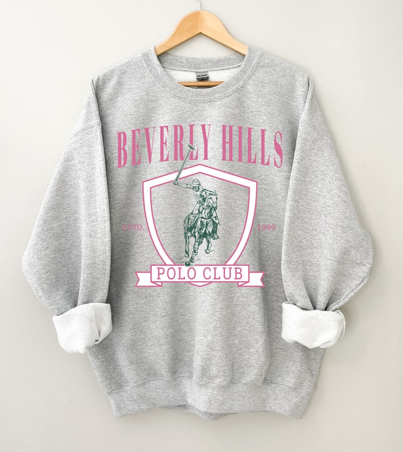 May include: Gray sweatshirt with a pink "Beverly Hills Polo Club" graphic featuring a polo player on a horse. The graphic is inside a shield with the years "Est'd 1990" and "1990" on either side.