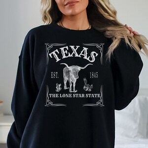 May include: Black sweatshirt with a white graphic featuring the word "TEXAS", a longhorn, cacti, and the text "EST. 1845" and "THE LONE STAR STATE". The design is centered on the front of the garment.
