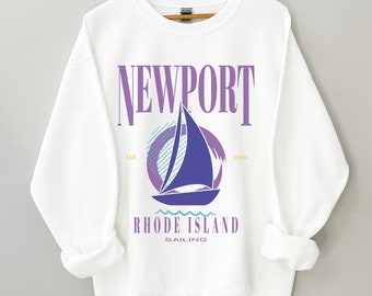 May include: White sweatshirt with a purple and blue graphic design of a sailboat with the text "NEWPORT" above the sailboat and "RHODE ISLAND SAILING" below the sailboat. The numbers "19" and "85" are on either side of the sailboat.