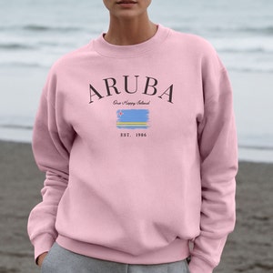 May include: A light pink sweatshirt with the word "ARUBA" in black arched text above a stylized Aruba flag and the text "One Happy Island" and "EST. 1986". The sweatshirt is a crew neck style.