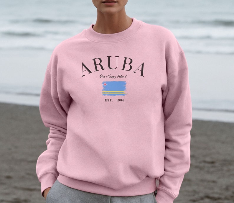 May include: A light pink sweatshirt with the word "ARUBA" in black arched text above a stylized Aruba flag and the text "One Happy Island" and "EST. 1986". The sweatshirt is a crew neck style.