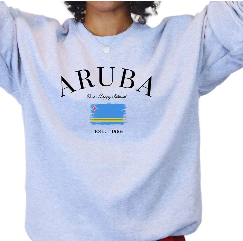 May include: Light gray sweatshirt with the word "ARUBA" in black, arched across the top. Below, the text "One Happy Island" and a stylized Aruba flag. The bottom reads "EST. 1986".
