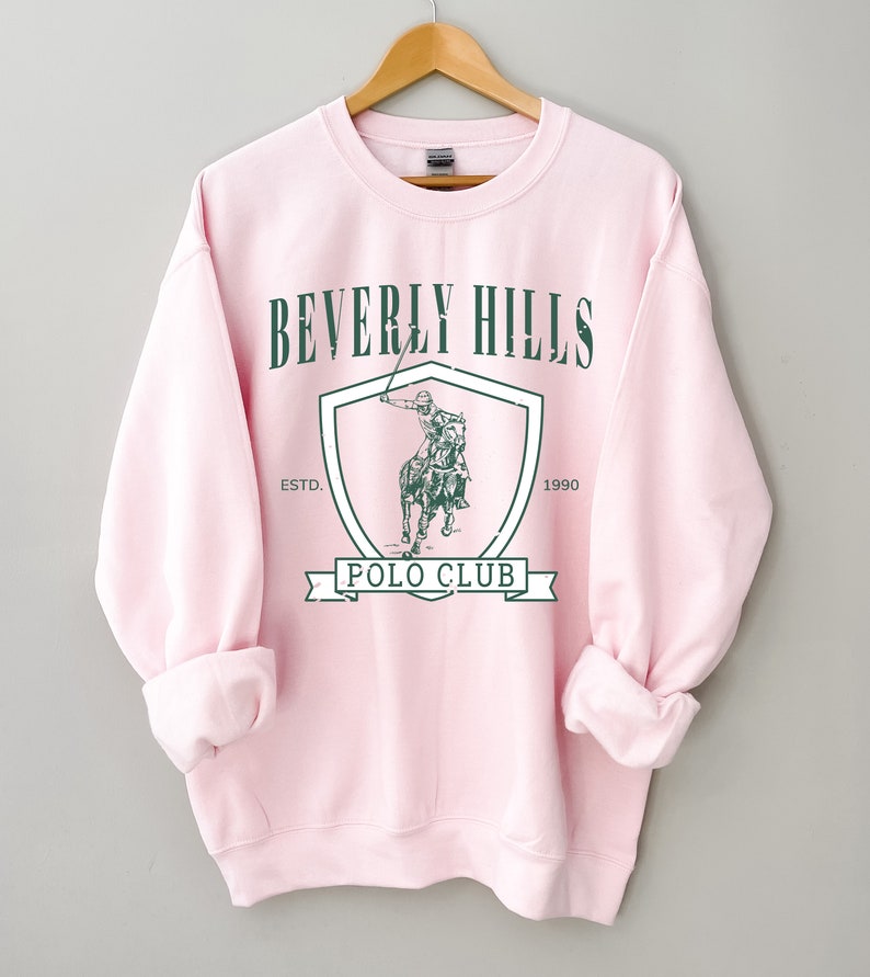 May include: Pink sweatshirt with a green graphic of a polo player on a horse inside a shield with the text "Beverly Hills Polo Club" and "Est. 1990".