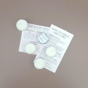 May include: Overhead shot of four white tea light candles, a small tin labeled "Dark Rose & Labdanum", and two informational cards. The cards have text including "How to Smell Like a Pro" and "The Icon Fragrance".