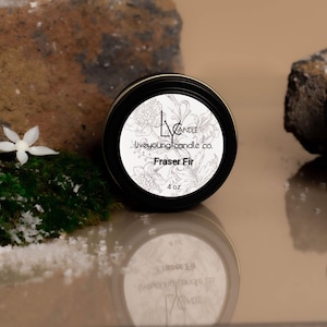 May include: A round black candle tin with a white label featuring the text "liveyoungcandle co." and "Fraser Fir." The label also includes a floral design and states "4 oz." The candle sits on a reflective surface.