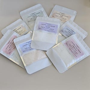 May include: A collection of eight white, resealable pouches containing laundry soap samples. Each pouch features a rectangular label with the text Laundry Soap Sample, a specific scent name, fragrance notes, and a net weight of 1 oz (28g).