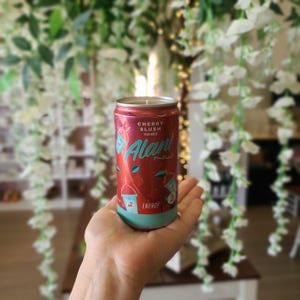 May include: A red and teal can of Alani Nu Cherry Slush flavored energy drink. The can features the brand name in teal script, cherry illustrations, and the word "Energy". The can is held in a hand.