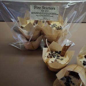 May include: A set of four fire starters in clear packaging. Each starter is shaped like a muffin, with a white wax base, topped with dark brown seeds and a cinnamon stick. The packaging includes text that reads "Fire Starters".