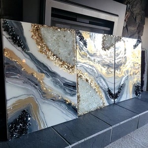 May include: A set of three abstract paintings with a white, gray, and gold color scheme. The paintings feature a swirling, marbled design with black and white glitter accents.