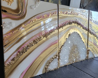 Puede incluir: Un conjunto de tres piezas de arte abstracto con un esquema de color blanco, rosa y dorado. La obra de arte presenta un diseño de remolino, similar al ágata, con acentos dorados y cristales blancos.