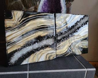Könnte beinhalten: Zwei abstrakte Harz-Kunstdrucke in Schwarz und Gold mit weißen und schwarzen Kristallen. Die Platten sind auf einer grauen Fliesenfläche platziert.
