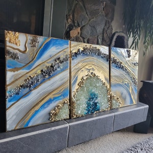 May include: Three abstract paintings on canvas with a blue, white, and gold color scheme. The paintings feature a swirling, agate-like design with gold accents.