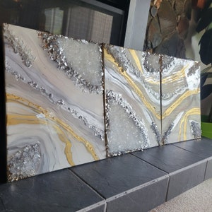 May include: Three abstract art panels with a white, gray, and gold color scheme. The panels feature a swirling, marbled design with a textured, glittery finish.