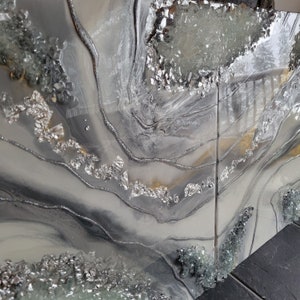 May include: A gray and white abstract painting with silver and gold accents. The painting features a swirling, flowing design with a textured surface. The painting is framed and ready to hang.