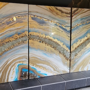 May include: A three-panel abstract art piece with gold, white, and blue colors. The artwork features a swirling, marbled design with embedded crystals.