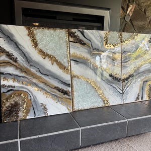 May include: A three-panel abstract art piece with a marbled design. The artwork features white, gray, and black swirls, accented with gold and crystal-like embellishments. The panels are displayed on a dark gray tiled surface.