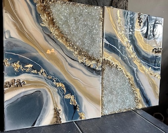 Puede incluir: Dos pinturas abstractas con un esquema de color azul, blanco y dorado. Las pinturas presentan un diseño de mármol en espiral con acentos de pan de oro.
