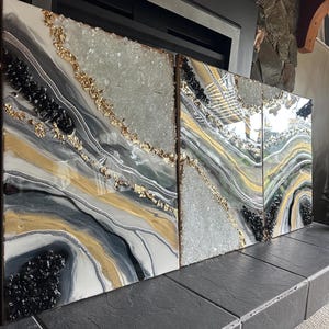 May include: Abstract wall art composed of three panels. Each panel features a swirling design in shades of gray, white, and gold, accented with black and clear crystal-like embellishments. Gold leaf adds a touch of luxury to the artwork.
