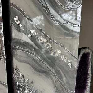 May include: A white and gray abstract resin art piece with silver glitter and silver leaf accents. The piece has a flowing, organic design.