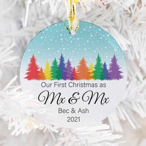 Puede incluir: Un adorno navideño redondo con fondo azul y una escena nevada. Presenta una fila de árboles de colores arcoíris y el texto "Our First Christmas as Mx & Mx Bec & Ash 2021".