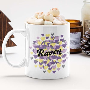 Puede incluir: Taza de cerámica blanca con un diseño de corazones morados y amarillos y el nombre "Raven" en texto negro.