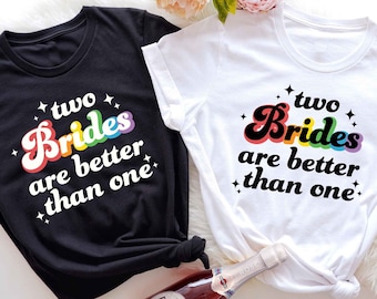 May include: Two black and white t-shirts with the text "two brides are better than one" in rainbow letters. The text is in a retro font style.