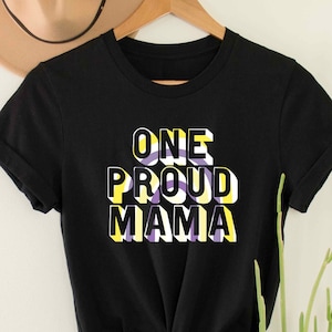Pode incluir: Camiseta preta com o texto "ONE PROUD MAMA" em letras amarelas, roxas e brancas. As letras estão em estilo 3D.