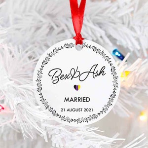 May include: White round bauble with a black wreath design, the text "Bex & Ash Married 21 August 2021" and a small heart with a purple, yellow, and white flag inside.