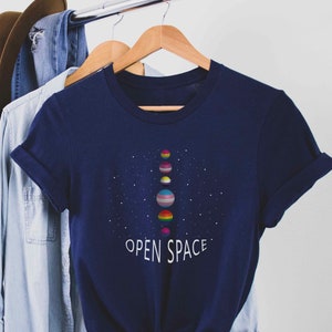 Puede incluir: Una camiseta azul marino con un gráfico de planetas en línea con un esquema de color arcoíris. El texto "OPEN SPACE" está impreso debajo de los planetas.