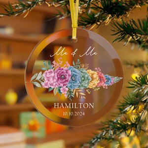 May include: A round glass bauble featuring a floral design in shades of pink, blue, and yellow. The bauble is inscribed with the text "Mr & Mr" and "HAMILTON 10.10.2024".