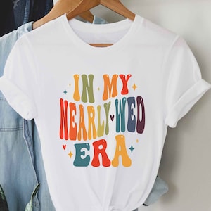 May include: White t-shirt with a rainbow-coloured graphic that says "In My Nearly-Wed Era".