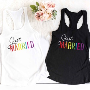以下が含まれることがあります： 白と黒のレーサーバックタンクトップ。筆記体で「Just Married」の文字が虹色で書かれています。白いふわふわした表面に、ピンクとピーチ色の花のブーケが添えられています。