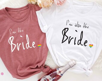 May include: Two white t-shirts with the text "I'm the Bride" and "I'm also the Bride" in black script. The shirts have a rainbow heart on the right side of the text. The shirt on the left is a dusty rose color.