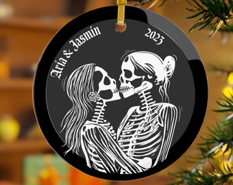 May include: Black and white glass ornament featuring a couple of skeletons kissing. The ornament is round and has a gold ribbon attached. The text "Aria & Jasmin 2023" is printed on the ornament.