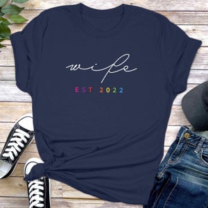 May include: A navy blue t-shirt with the word "wife" written in white cursive script and the text "EST 2022" in rainbow colors.