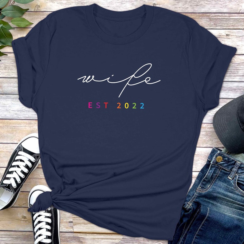May include: A navy blue t-shirt with the word "wife" written in white cursive script and the text "EST 2022" in rainbow colors.