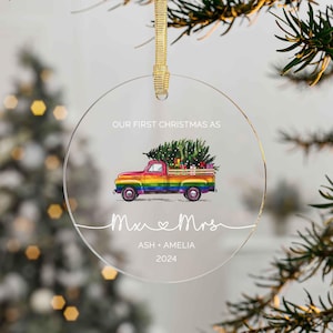 Puede incluir: Un adorno redondo de vidrio transparente con una camioneta de recogida de color arcoíris que lleva un árbol de Navidad. El texto "Our First Christmas as Mr & Mrs Ash + Amelia 2024" está impreso en el adorno.