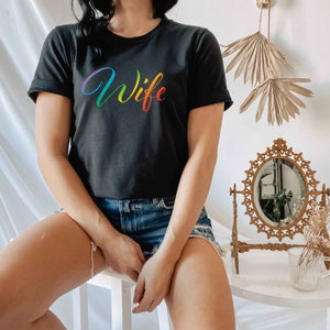 May include: A black t-shirt with a rainbow-colored "Wife" graphic printed on the front. The shirt is being worn by a person with denim shorts.