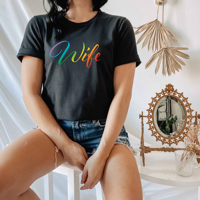 May include: A black t-shirt with a rainbow-colored "Wife" graphic printed on the front. The shirt is being worn by a person with denim shorts.