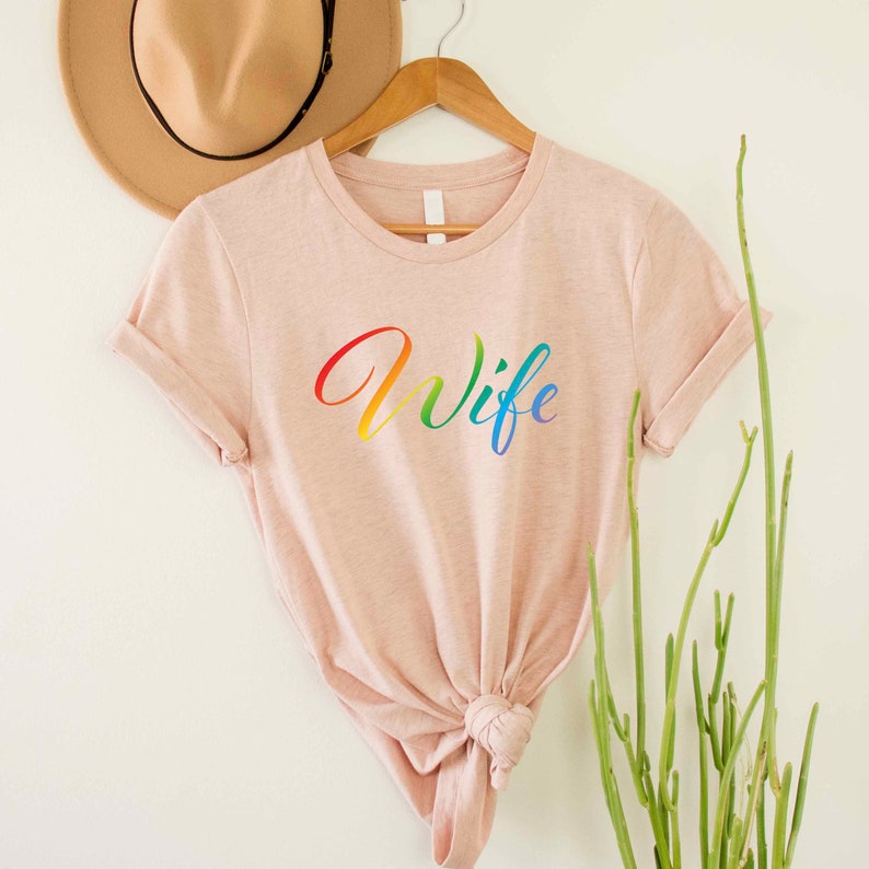 May include: A light pink t-shirt with the word "Wife" written in rainbow colors.