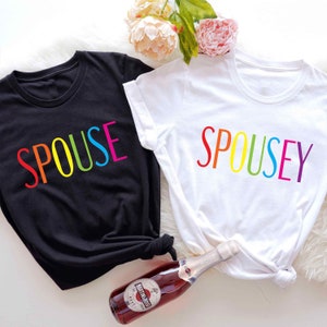 May include: Two t-shirts with the words "SPOUSE" and "SPOUSEY" printed in rainbow colors. The shirts are black and white and are laid out on a white surface.