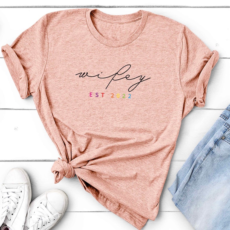 May include: A light pink t-shirt with the word "wifey" written in black cursive script. Below the word is the text "EST 2022" in rainbow colors.