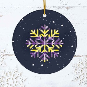 May include: A round, blue and white ceramic ornament with a yellow and purple snowflake design. The snowflake is a stylized design with stripes.