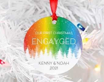 May include: A round, rainbow-colored Christmas ornament with a white silhouette of a forest and the text "Our First Christmas Engaged Kenny & Noah 2021".