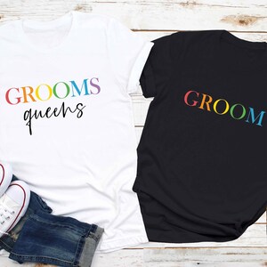 May include: Two white t-shirts with rainbow text. One shirt says "GROOMS queens" and the other says "GROOM".