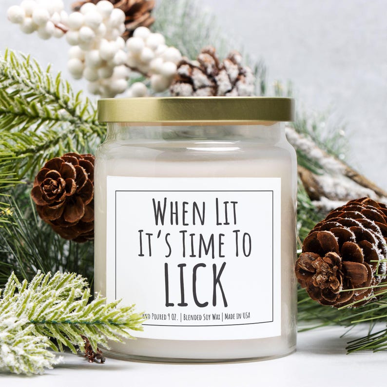 Puede incluir: Una vela en tarro de cristal transparente con tapa dorada. La vela tiene una etiqueta blanca con el texto "WHEN LIT IT'S TIME TO LICK". La vela est&aacute; rodeada de agujas de pino, pi&ntilde;as y bayas blancas. Hecho en EE. UU.