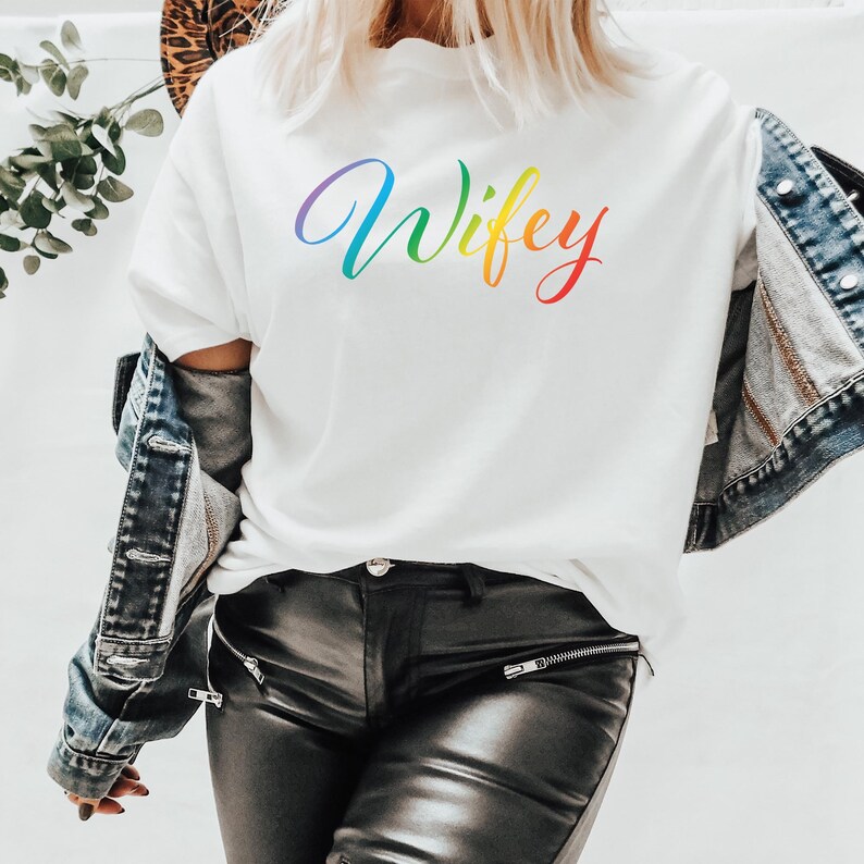 May include: White t-shirt with the word "Wifey" written in rainbow colors.